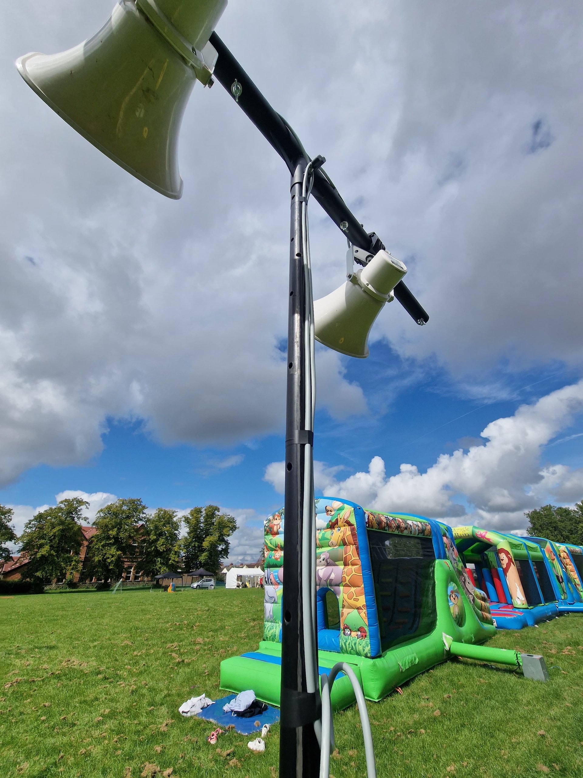 Outdoor event setup with mounted loudspeakers and inflatable attractions on a grassy field under a cloudy sky.