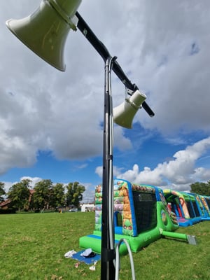 Outdoor event setup with mounted loudspeakers and inflatable attractions on a grassy field under a cloudy sky.