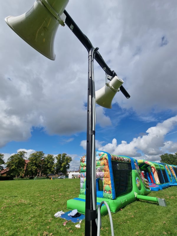 Outdoor event setup with mounted loudspeakers and inflatable attractions on a grassy field under a cloudy sky.