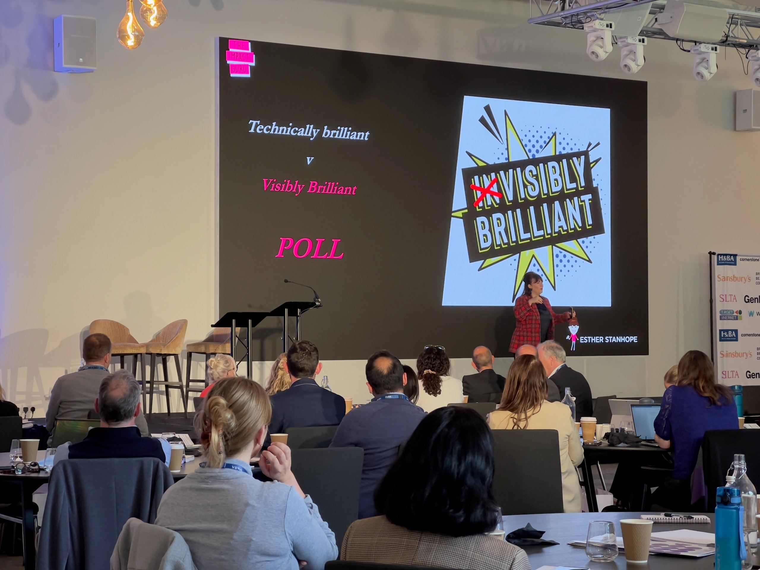 Speaker presenting on stage at a corporate conference, with audience seated at round tables and a large display screen behind them.