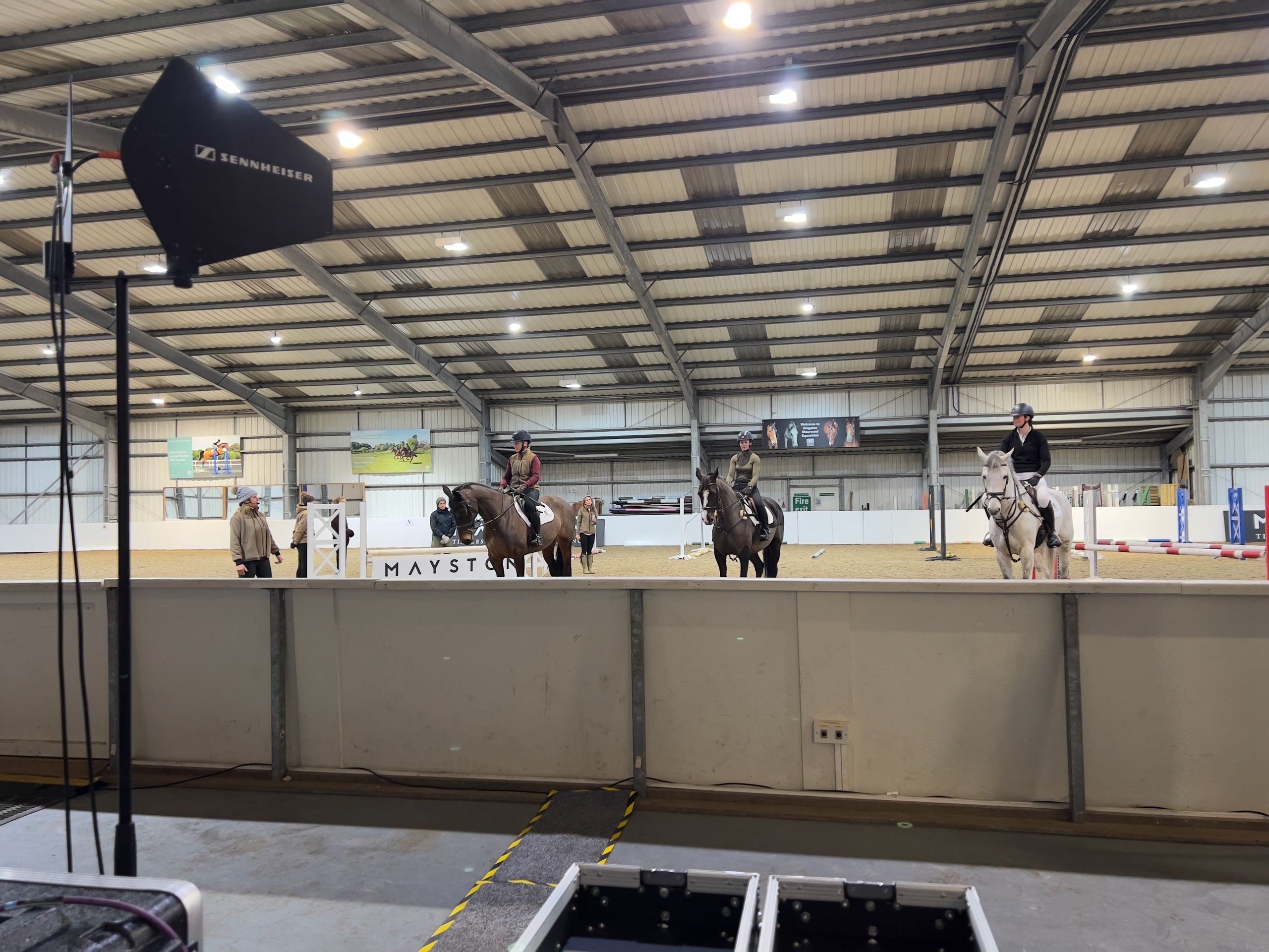 Indoor equestrian event setup with riders practicing under arena lighting, featuring prominent sound equipment in the foreground.