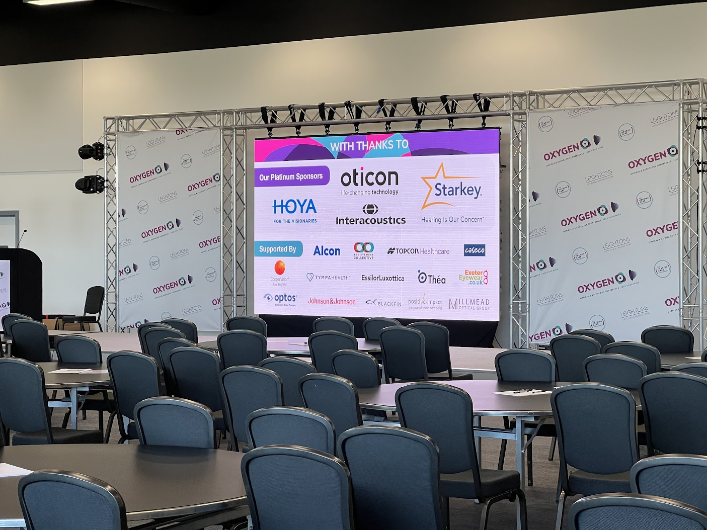 Conference room setup with staging, sponsor banners displayed on a large screen, and round tables with chairs arranged for attendees.