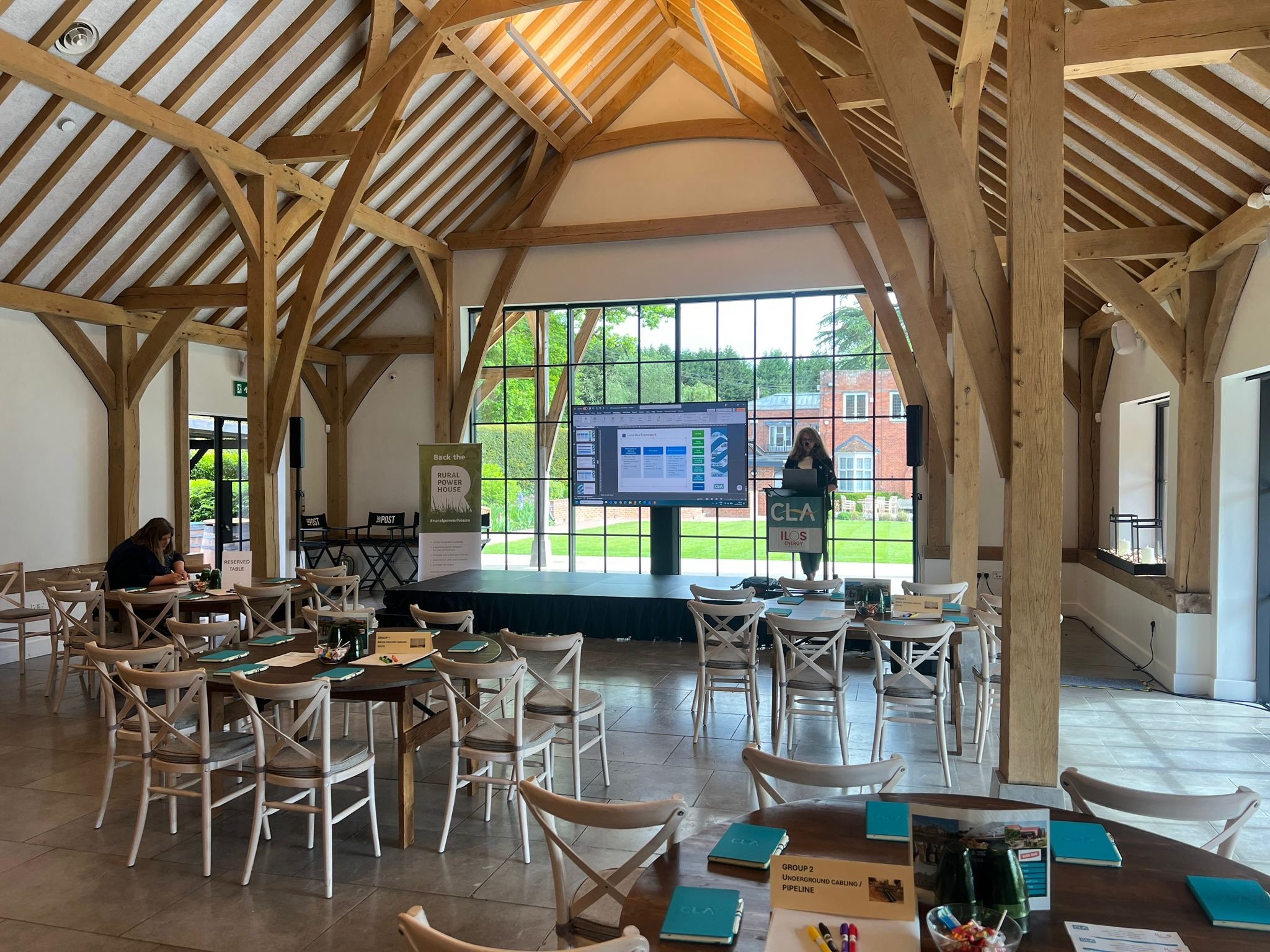 Speaker presenting on stage at a corporate conference, with audience seating and active sound and lighting setup in a wooden beam venue.