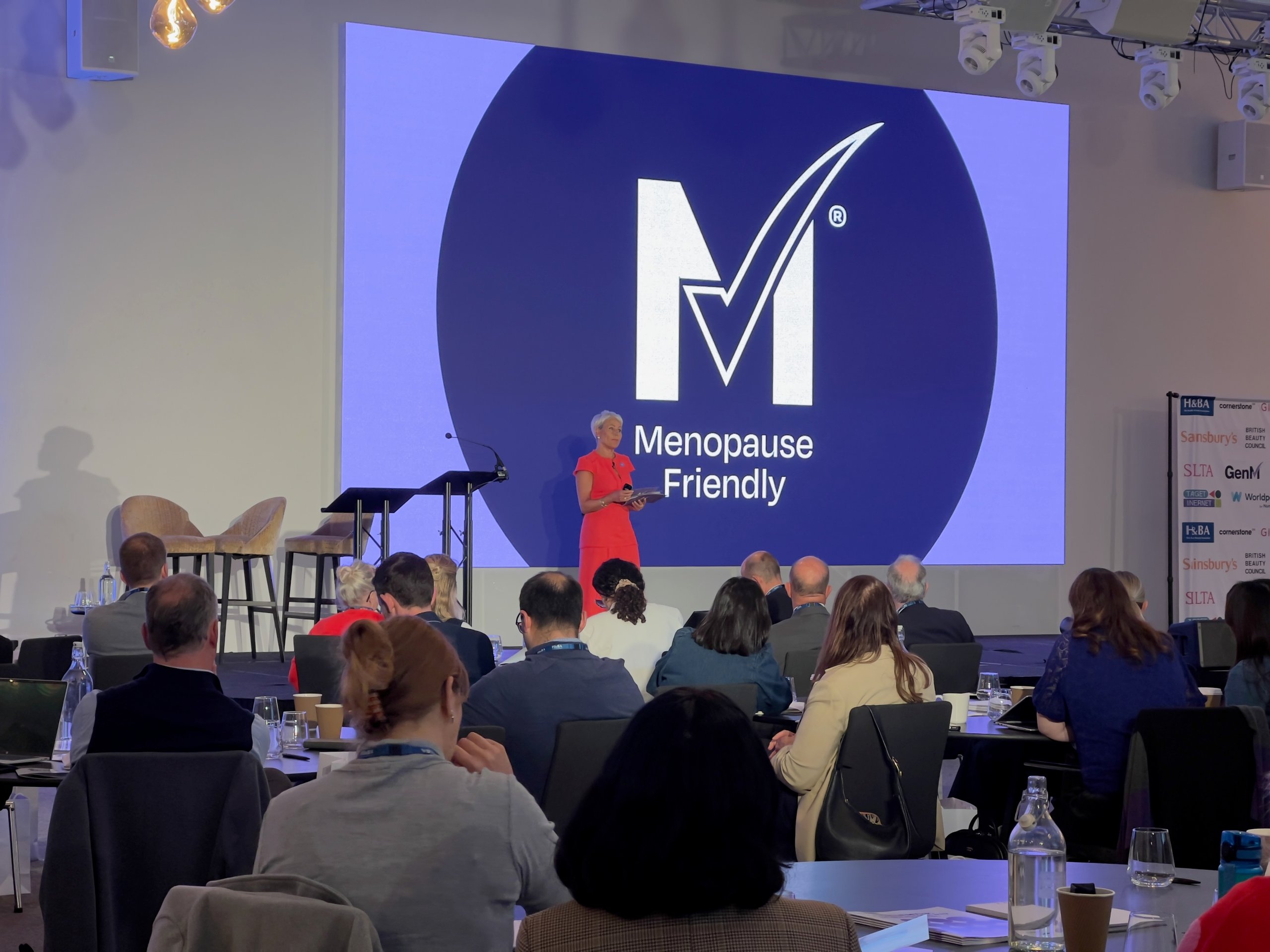 Speaker presenting at a corporate conference on menopause, featuring professional staging, lighting, and large video screen setup.