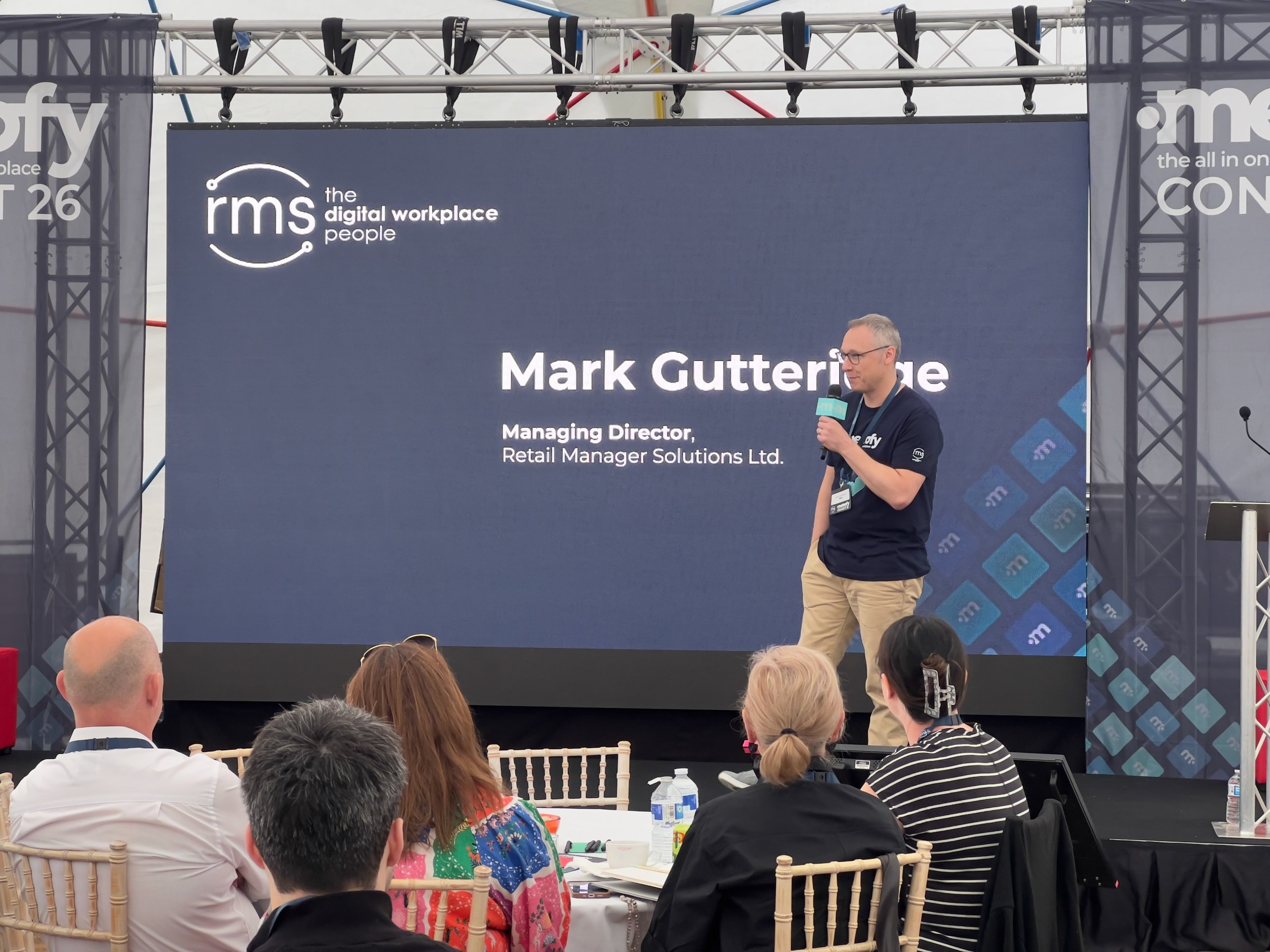 Speaker presenting on stage at corporate conference, with AV setup including backdrop screen and audience seated in foreground.