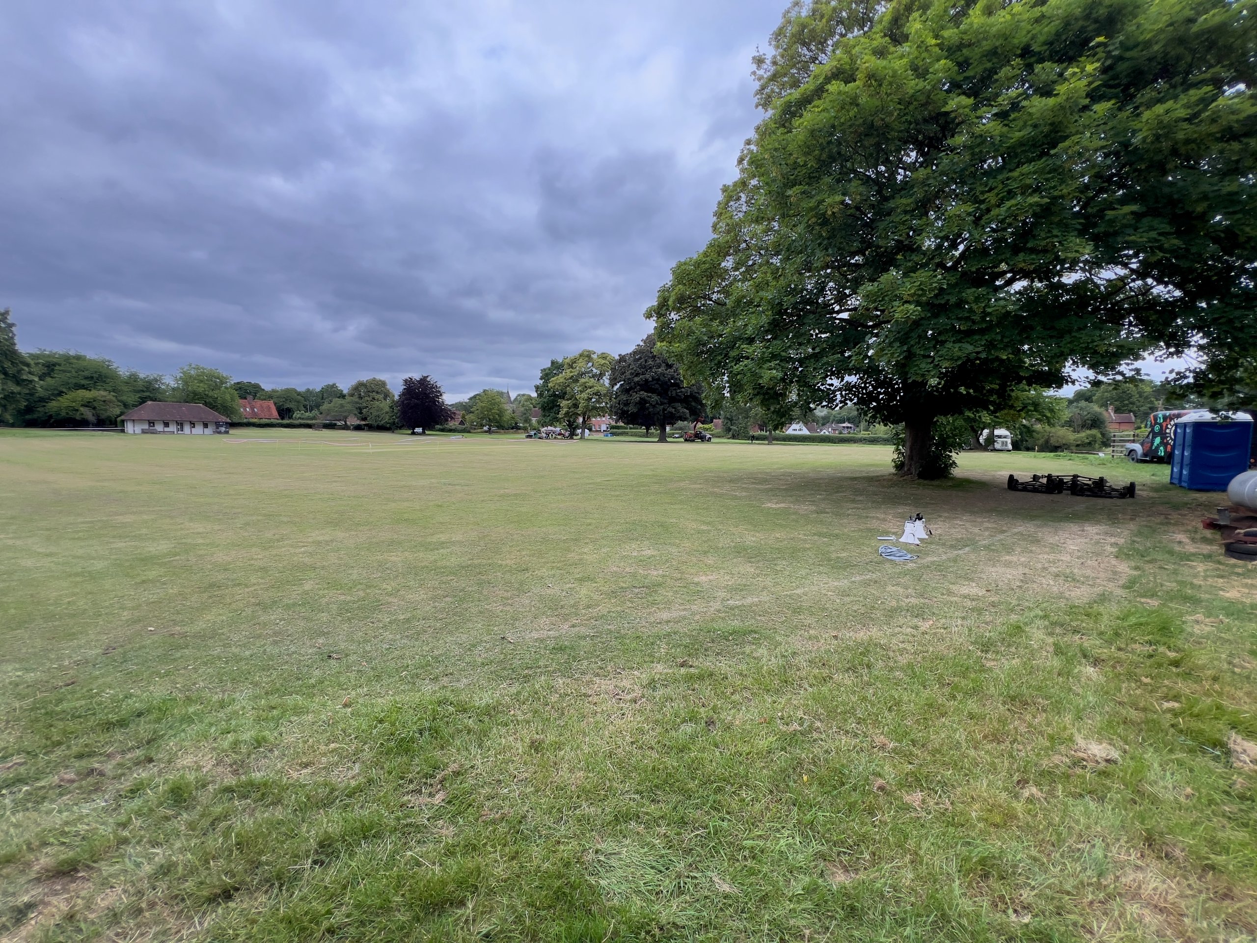 Expansive outdoor field on a cloudy day, with scattered AV equipment pieces under a large tree, suggesting event setup preparation.