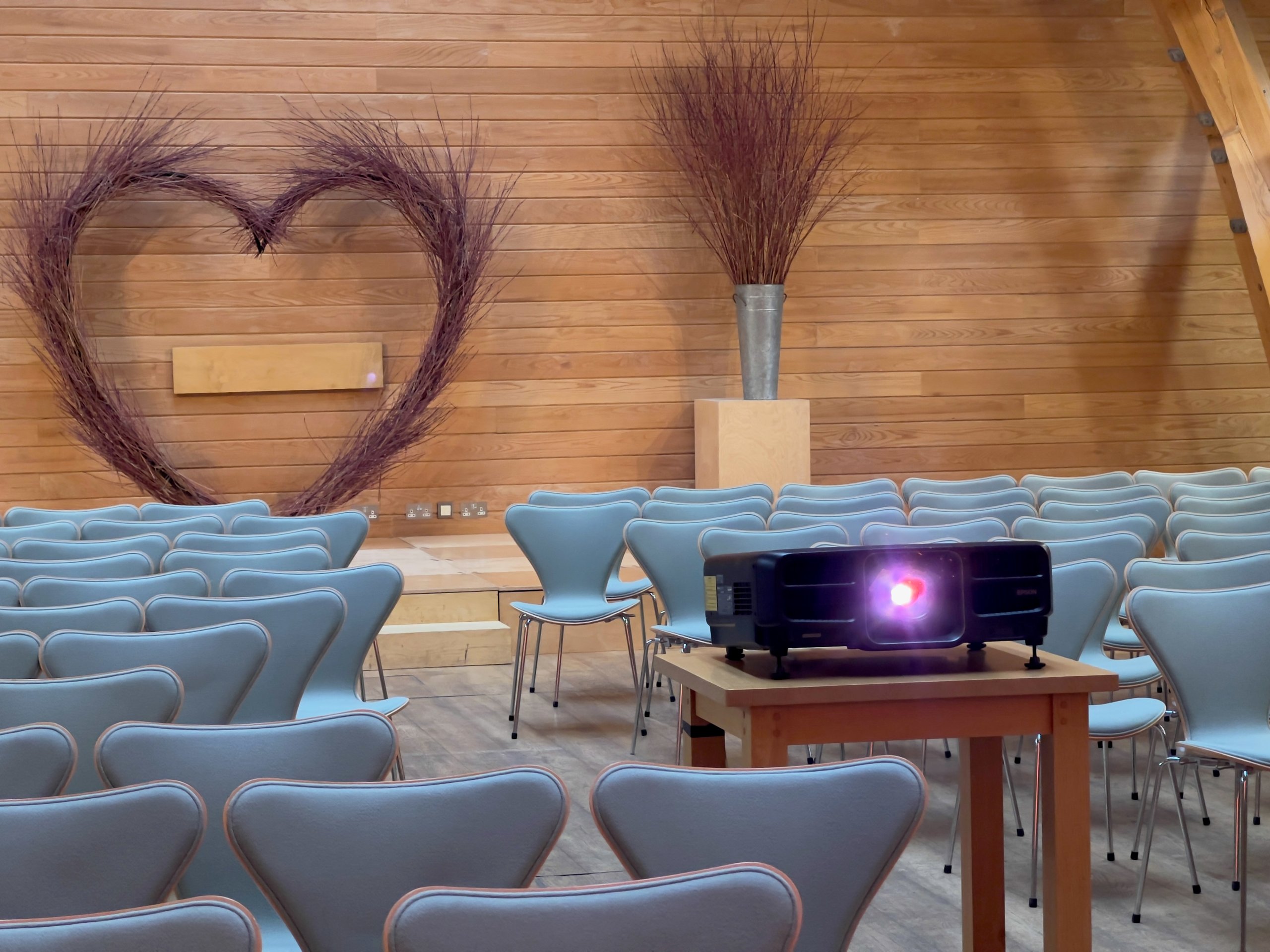 Projector setup in a wooden reception hall with decorative heart-shaped twig design, rows of chairs for a corporate event.