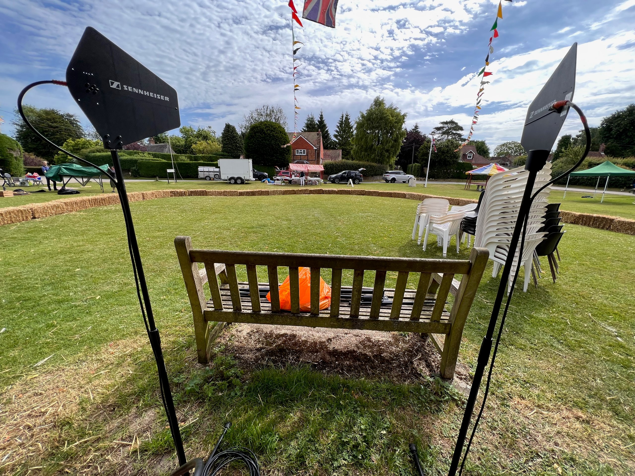 Outdoor event setup with Sennheiser sound equipment, a wooden bench, stacked chairs, and colorful flags on a grassy field.