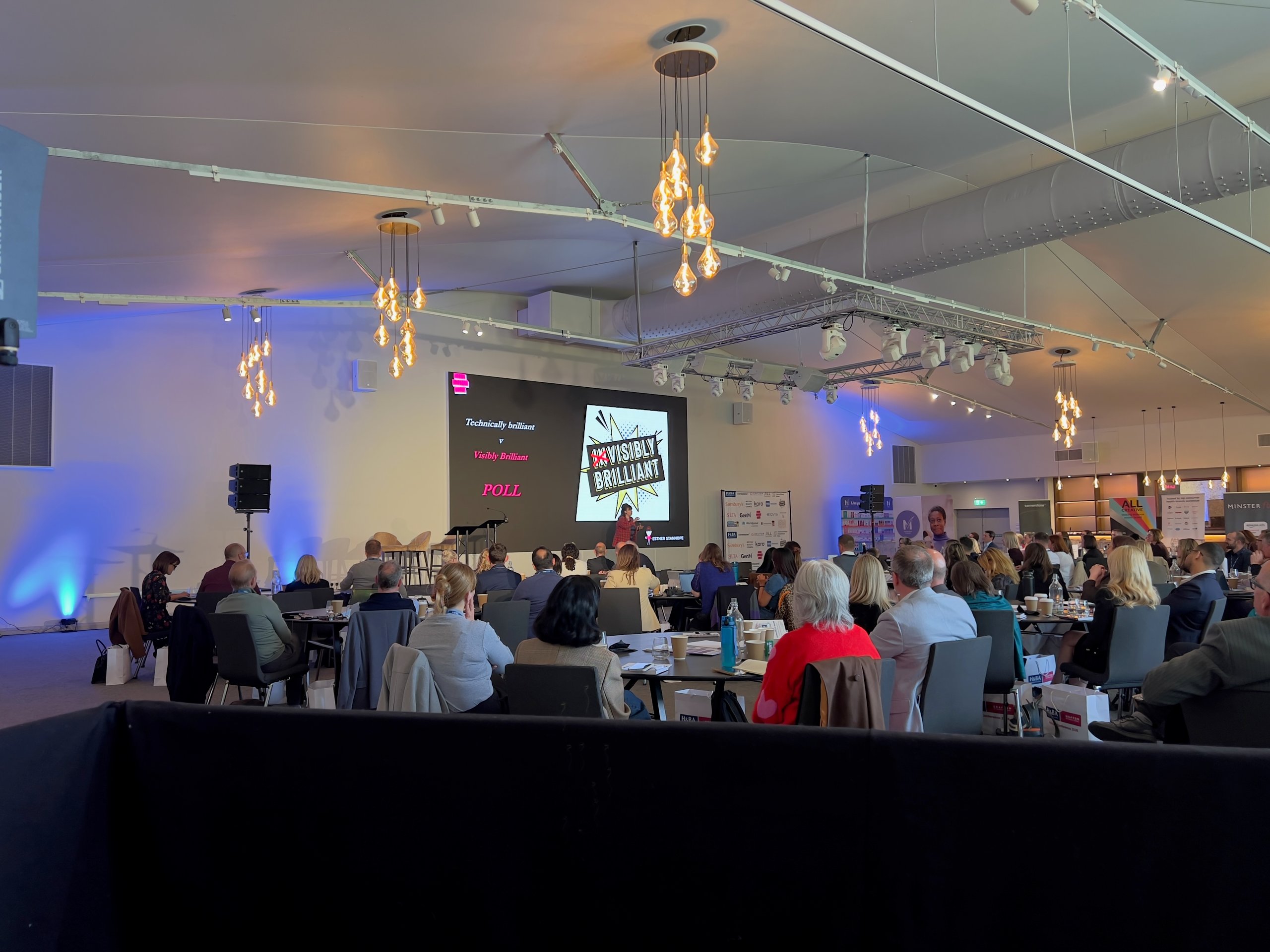 Delegates seated at a conference venue watching a presentation on a projector screen, with professional stage lighting.