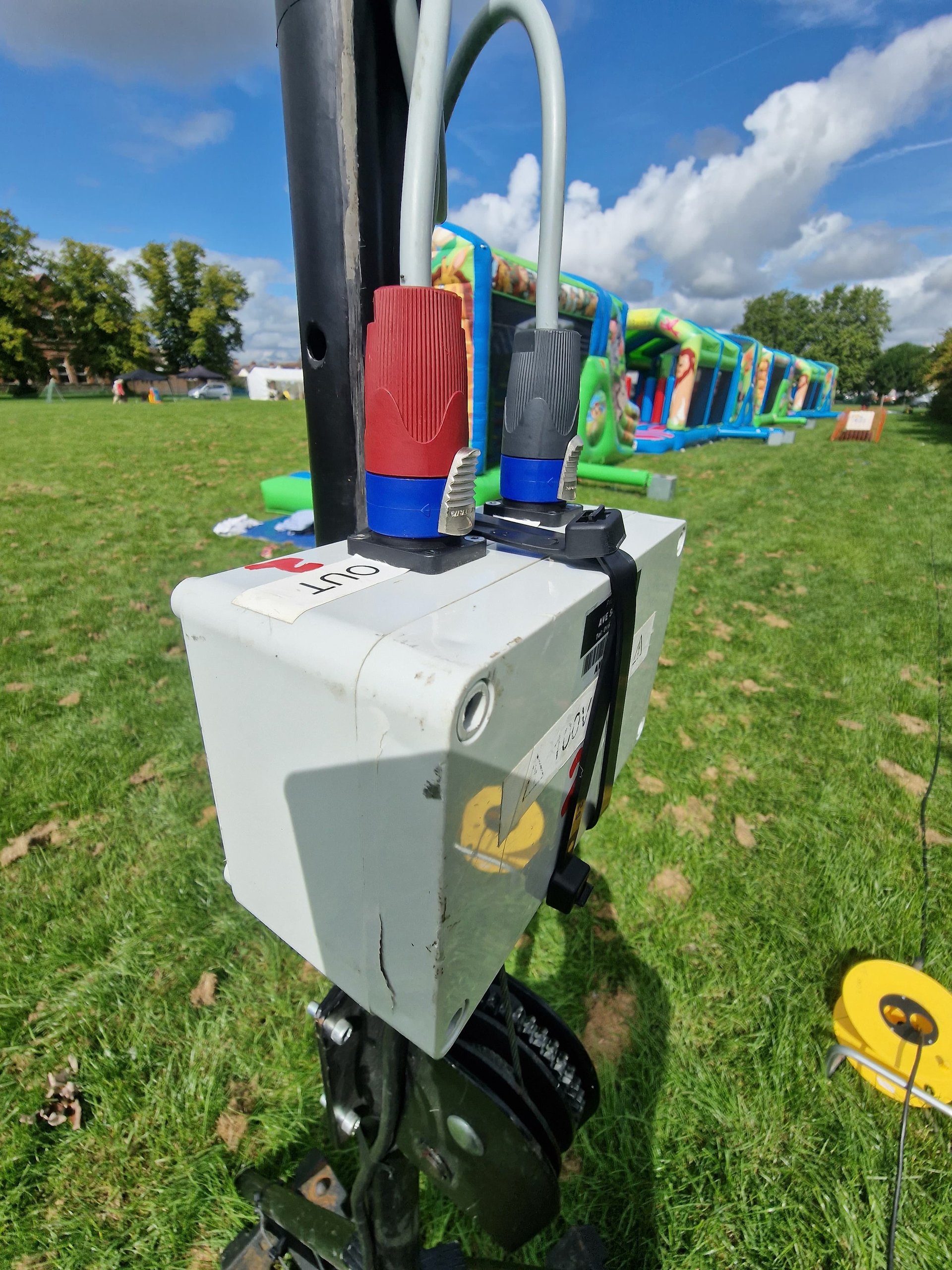 Outdoor festival setup with power distribution box and cables on a grassy field, inflatable attractions in the background.