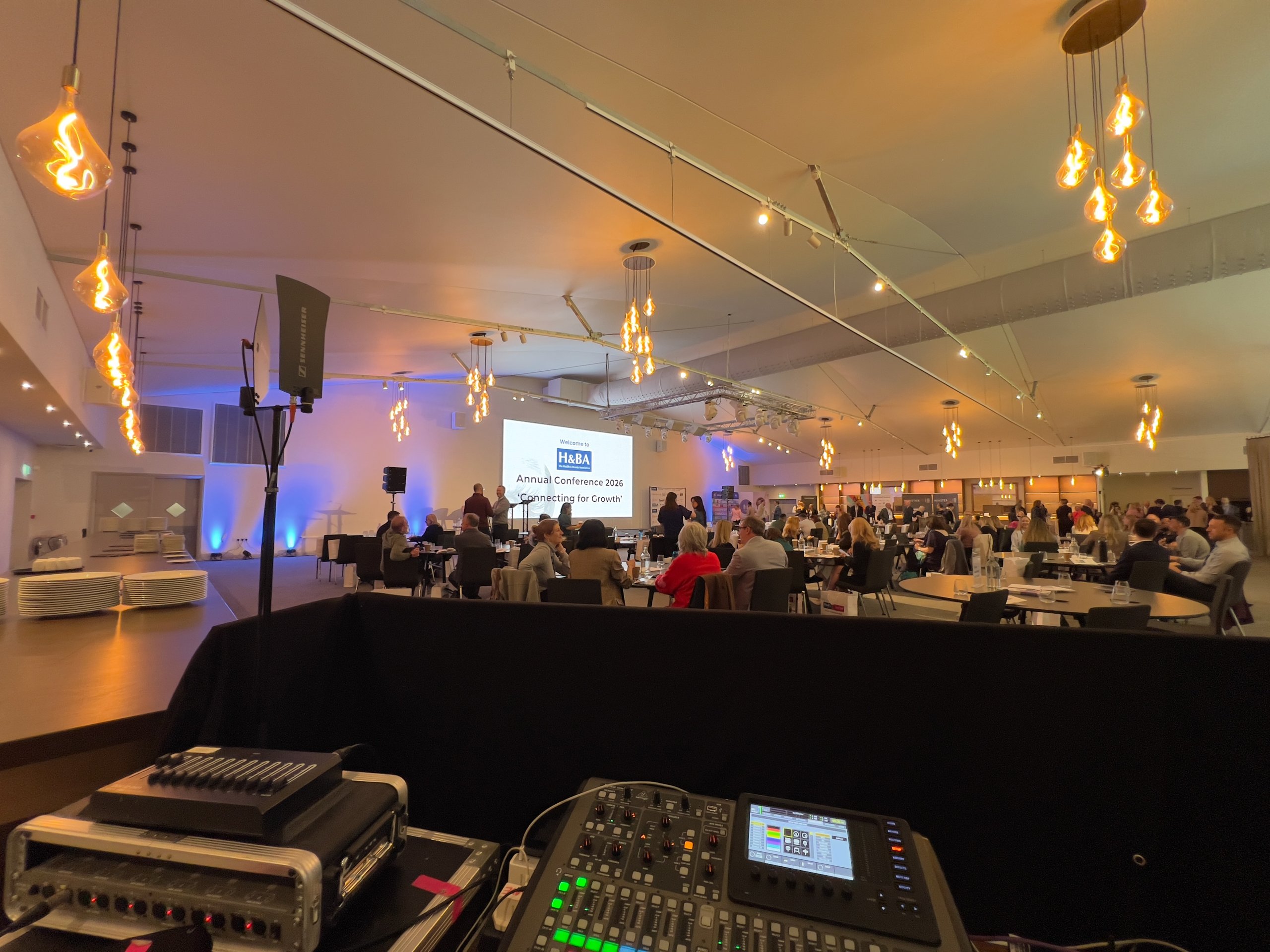 Corporate conference room with staging, lighting, and audio equipment. Guests seated at tables, facing a presentation screen.
