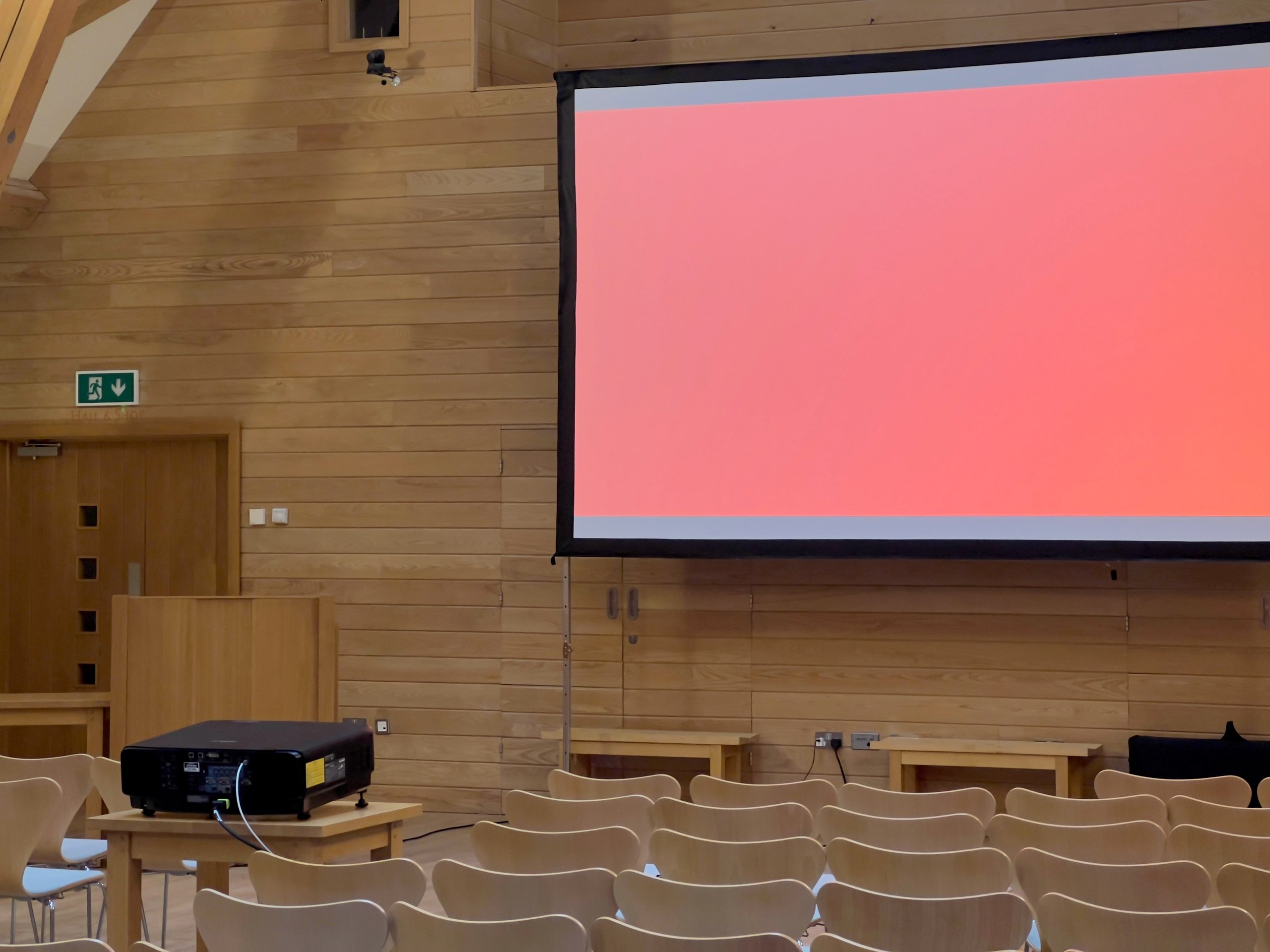 Conference room setup with a large screen displaying a red background, a projector on a table, and rows of empty chairs.