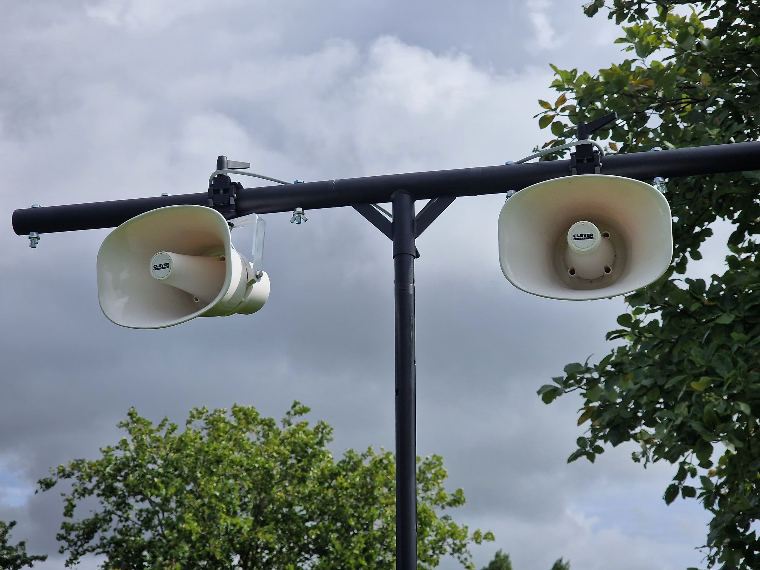 Mounted outdoor PA speakers on a metal stand amidst greenery and cloudy skies, ideal for open-air events and announcements.