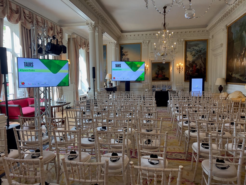 Conference setup in an elegant room featuring rows of chairs, stage lighting, and AV screens displaying Spain Talks.