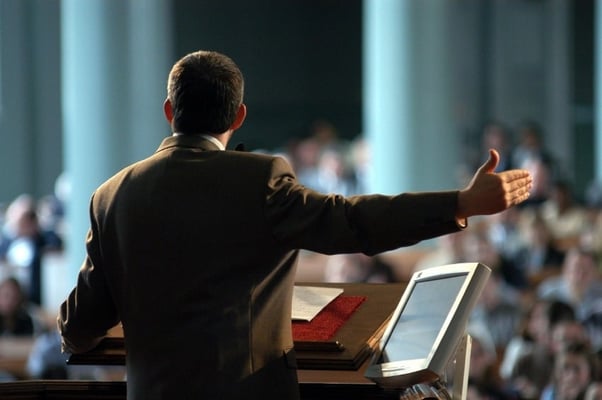 Speaker at a corporate conference using AV equipment, addressing an audience from a podium with a digital screen.