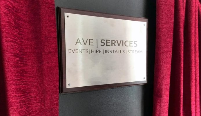 AVE Services metal sign with red curtains, highlighting events, AV hire, installations, and streaming services.