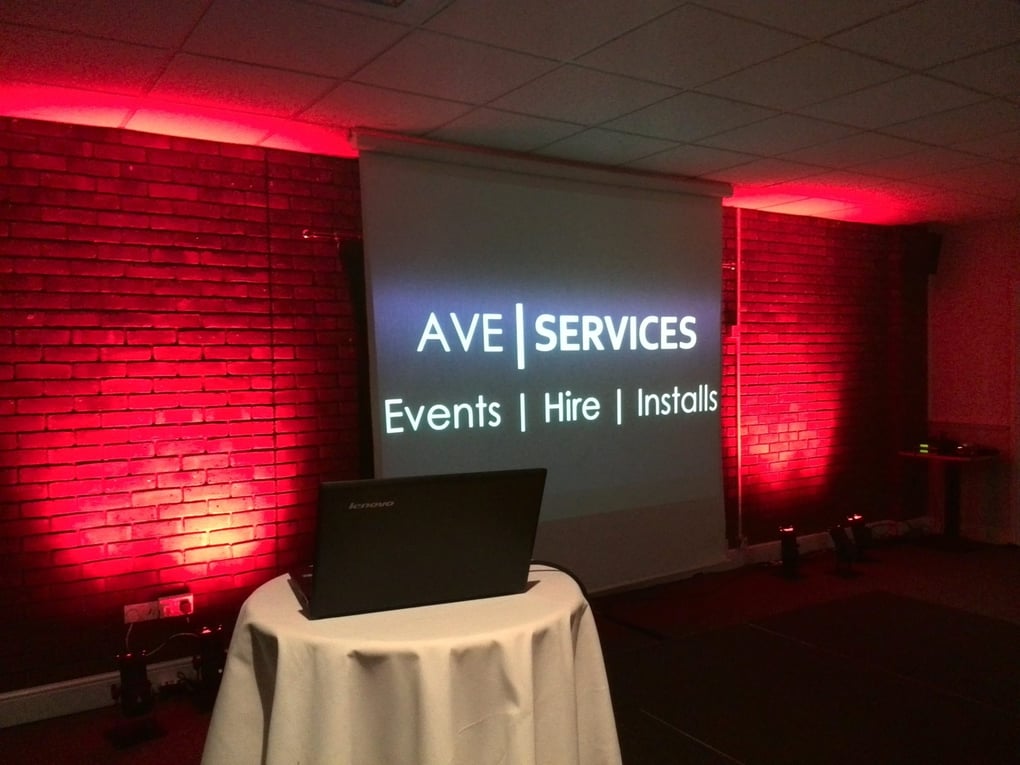 Projector screen displays AVE Services with red ambient lighting on brick walls, set up for a corporate event.