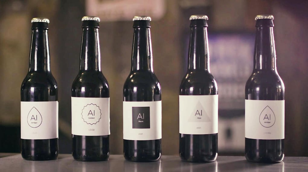 Five black beer bottles with minimalist white labels displaying different versions, lined up on a stainless steel counter.