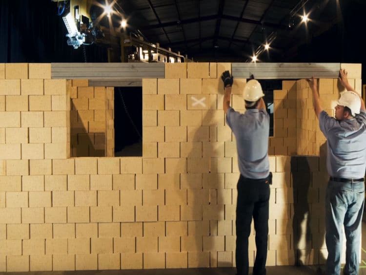 Two technicians in safety gear constructing a stage backdrop with lighting equipment overhead in an indoor event setting.