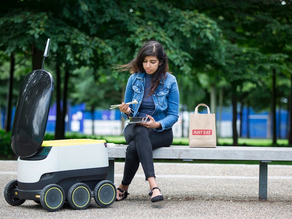 Woman seated on outdoor bench eating from takeout container beside a delivery robot and a Just Eat bag in a park setting.