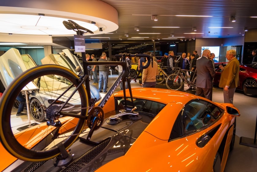 Exhibition featuring luxury cars and bicycles in a showroom setup; attendees engaging in discussion under ambient lighting.