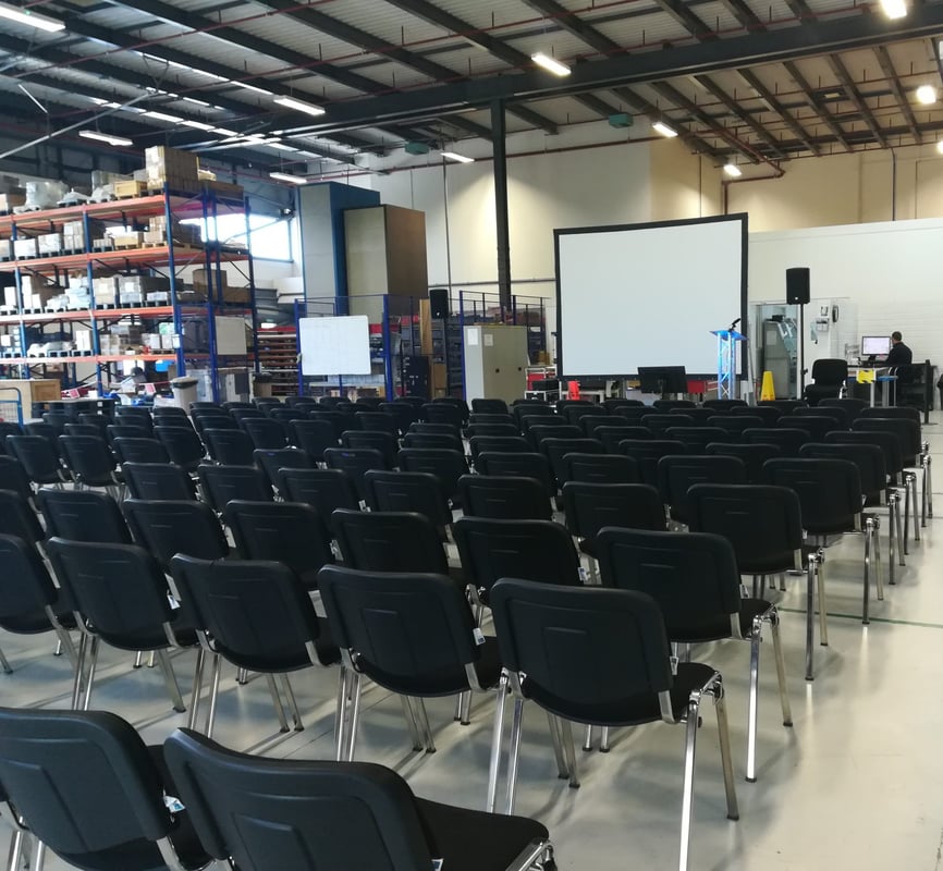 Rows of black chairs face a large projection screen and sound equipment in a warehouse setup for a corporate conference.