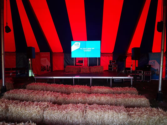 Staging setup in a red and blue tent for All Staff Away Day with hay bale seating and AV equipment.