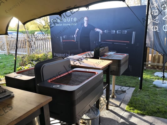 Outdoor event setup featuring Everdure grills under a tent, with promotional signage in the background.