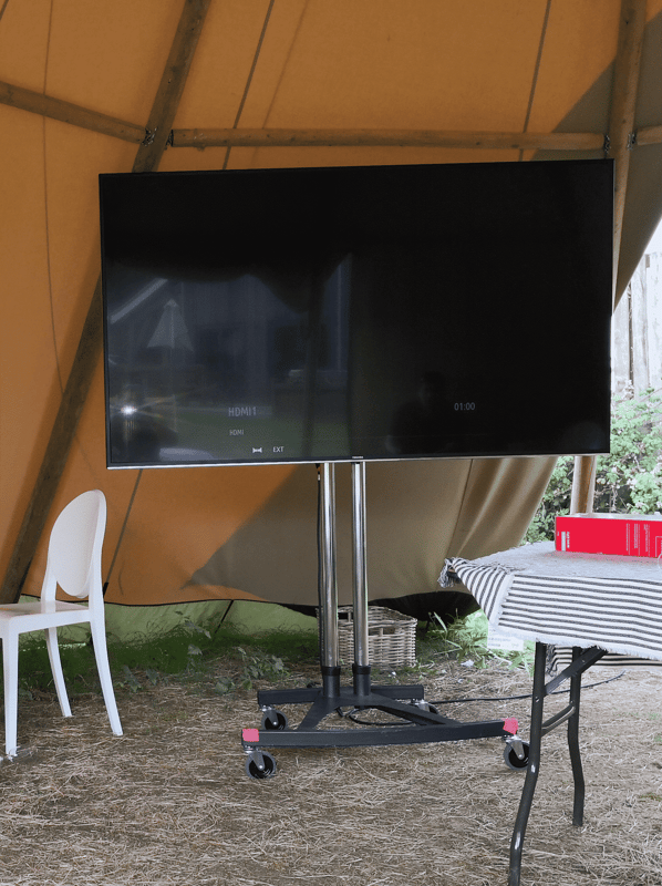 Large monitor on a mobile stand under a tent, set up for an outdoor event, alongside a table and chair. Ideal for presentations.