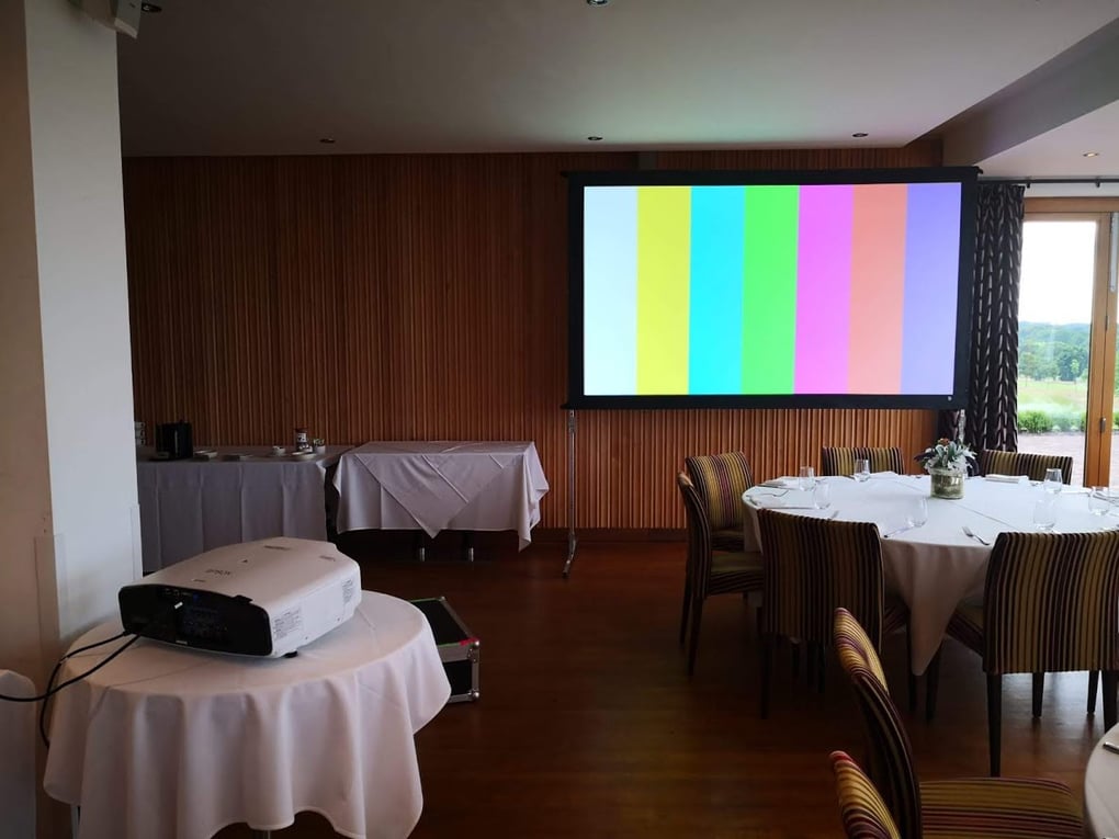 Projector setup in a conference room with a colorful test pattern on the screen, round tables ready for an event.