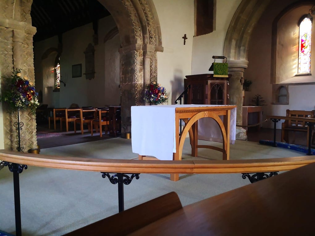 Church altar setup with white cloth in a historic venue, flanked by floral displays and wooden pews in the background.