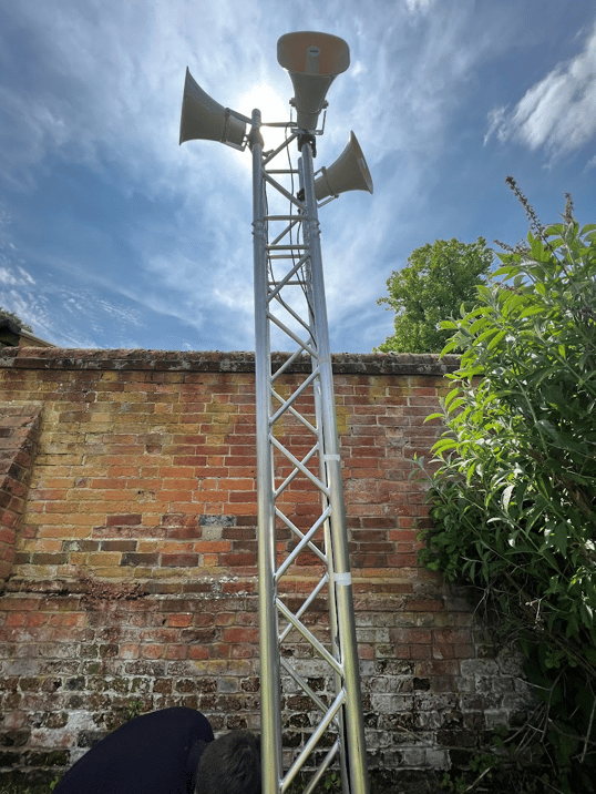 Steel truss with mounted loudspeakers set up outdoors against a brick wall, under a clear sky for an outdoor event.