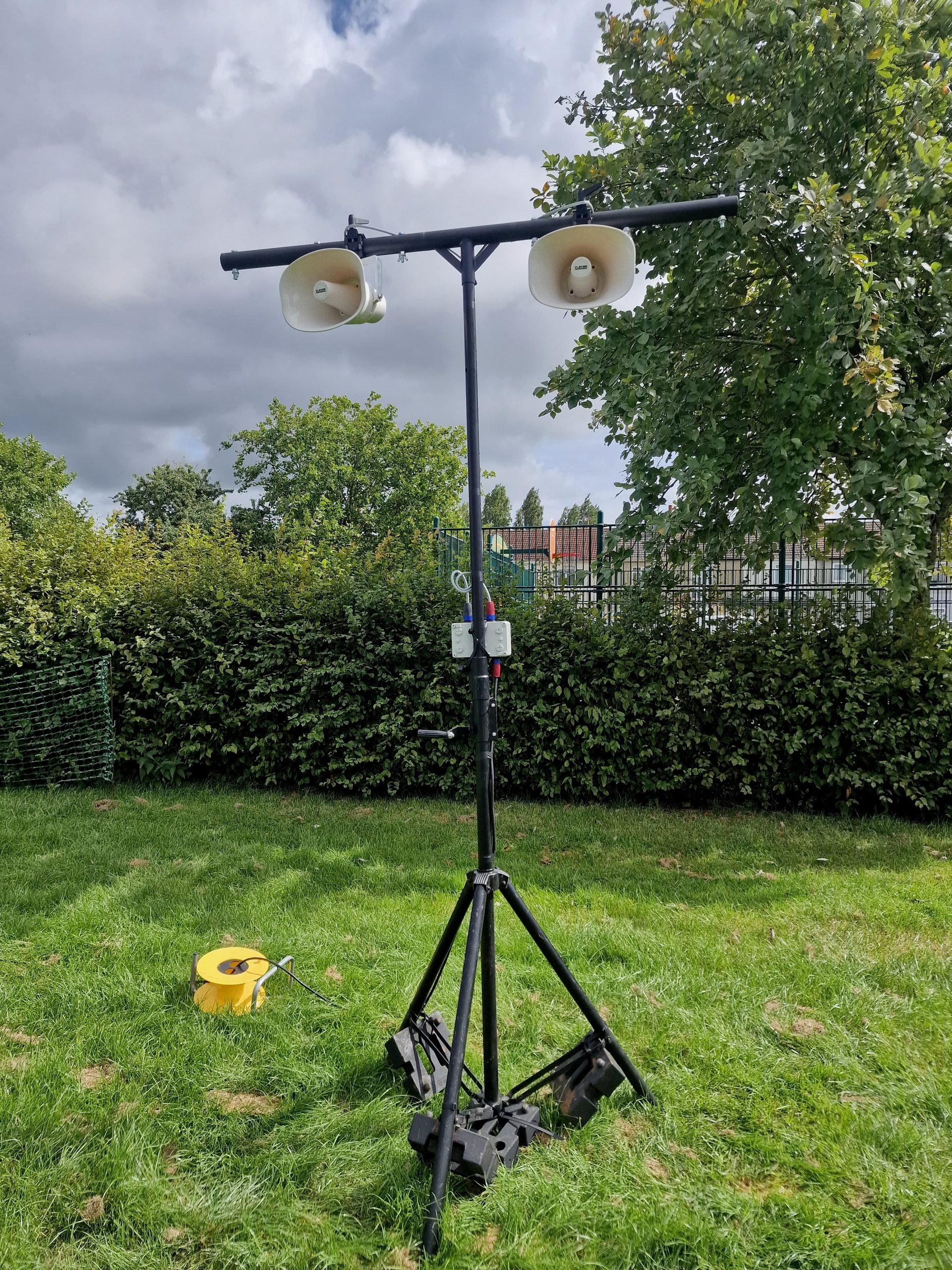 Outdoor PA speakers on a tripod stand, set up for event sound coverage in a garden setting.