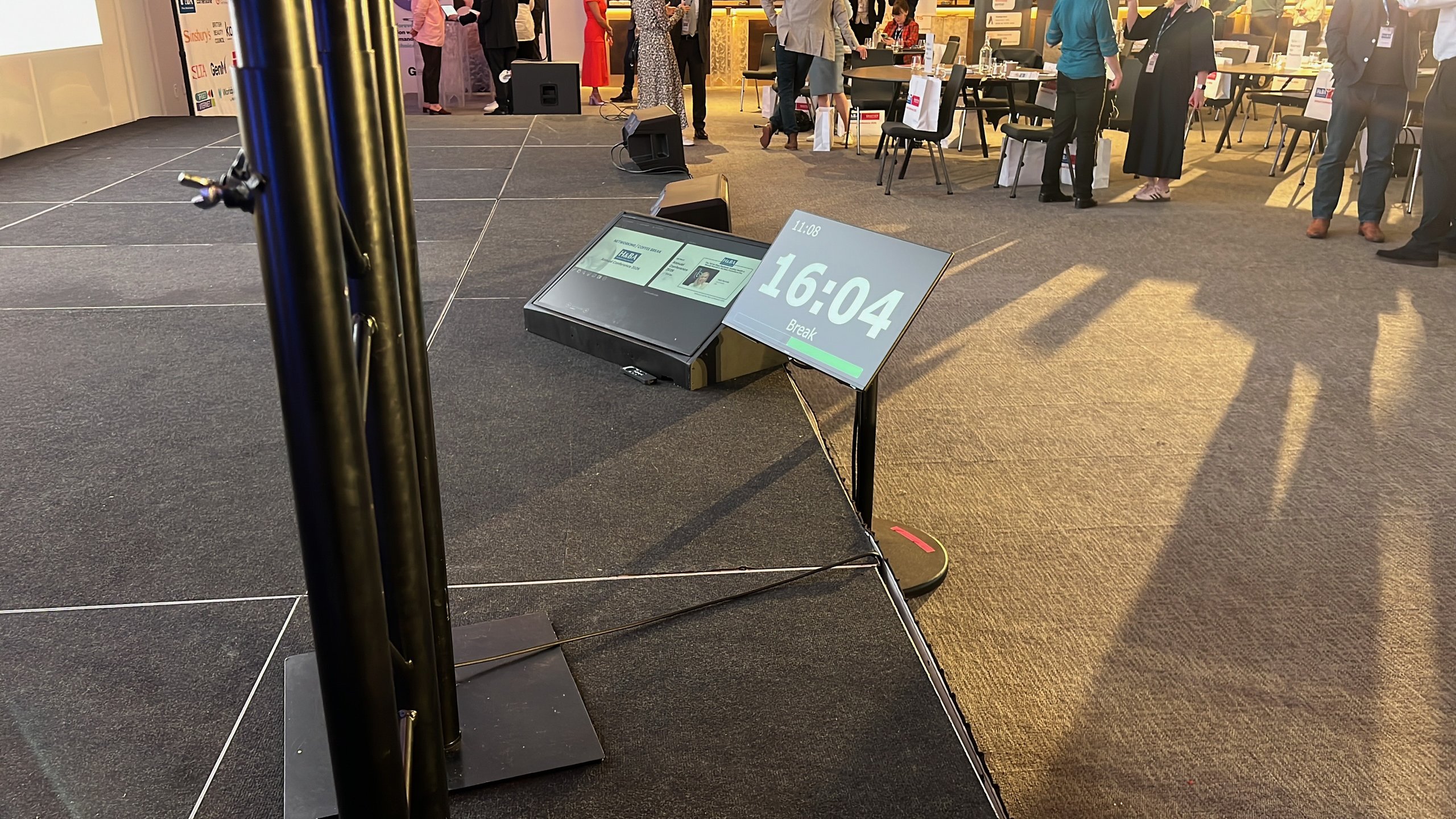 Stage with teleprompter and sound equipment setup for a corporate conference, attendees gather in the background during a break.