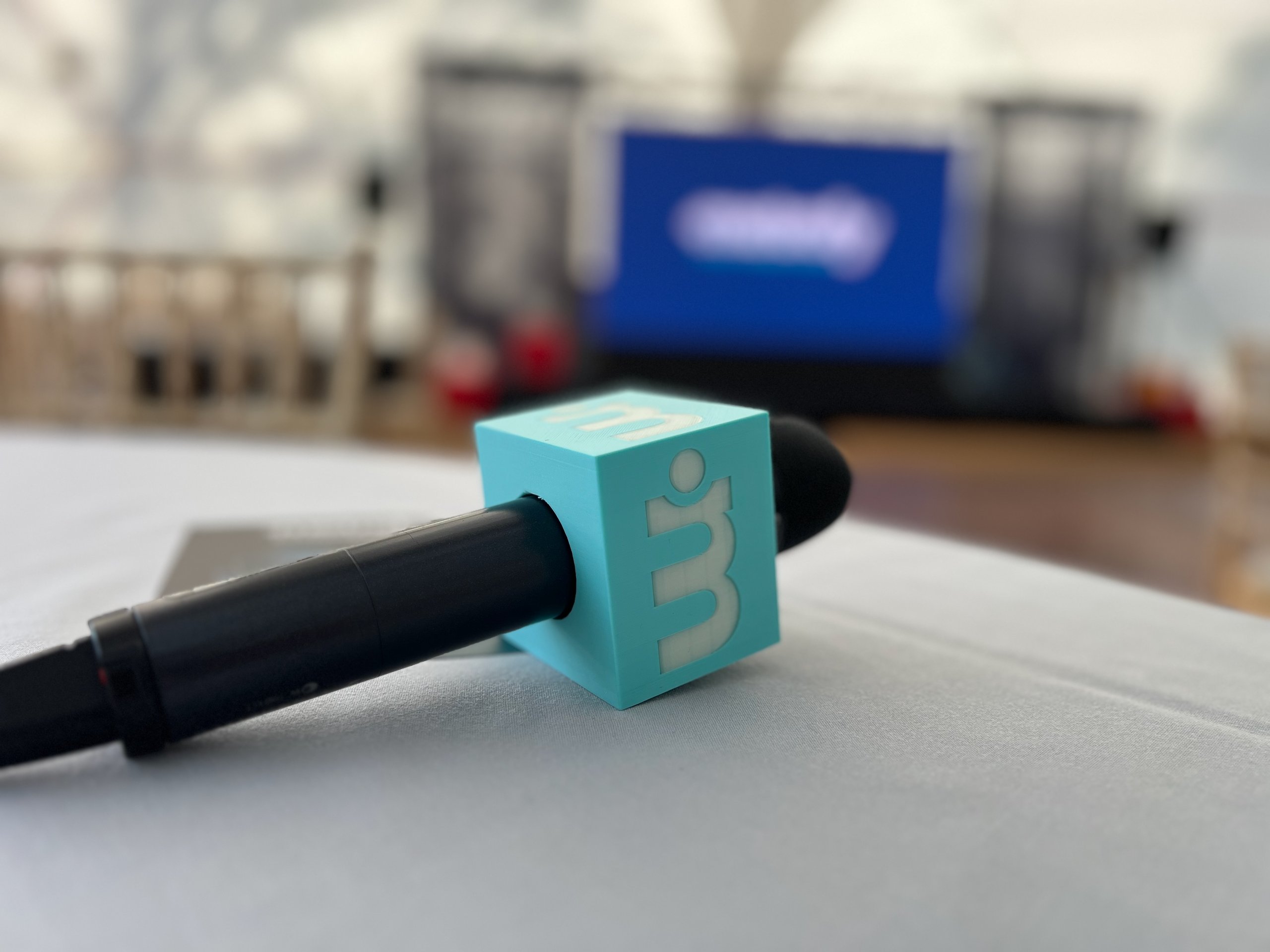 Microphone with logo on a table at a corporate event, conference stage and projection screen blurred in the background.