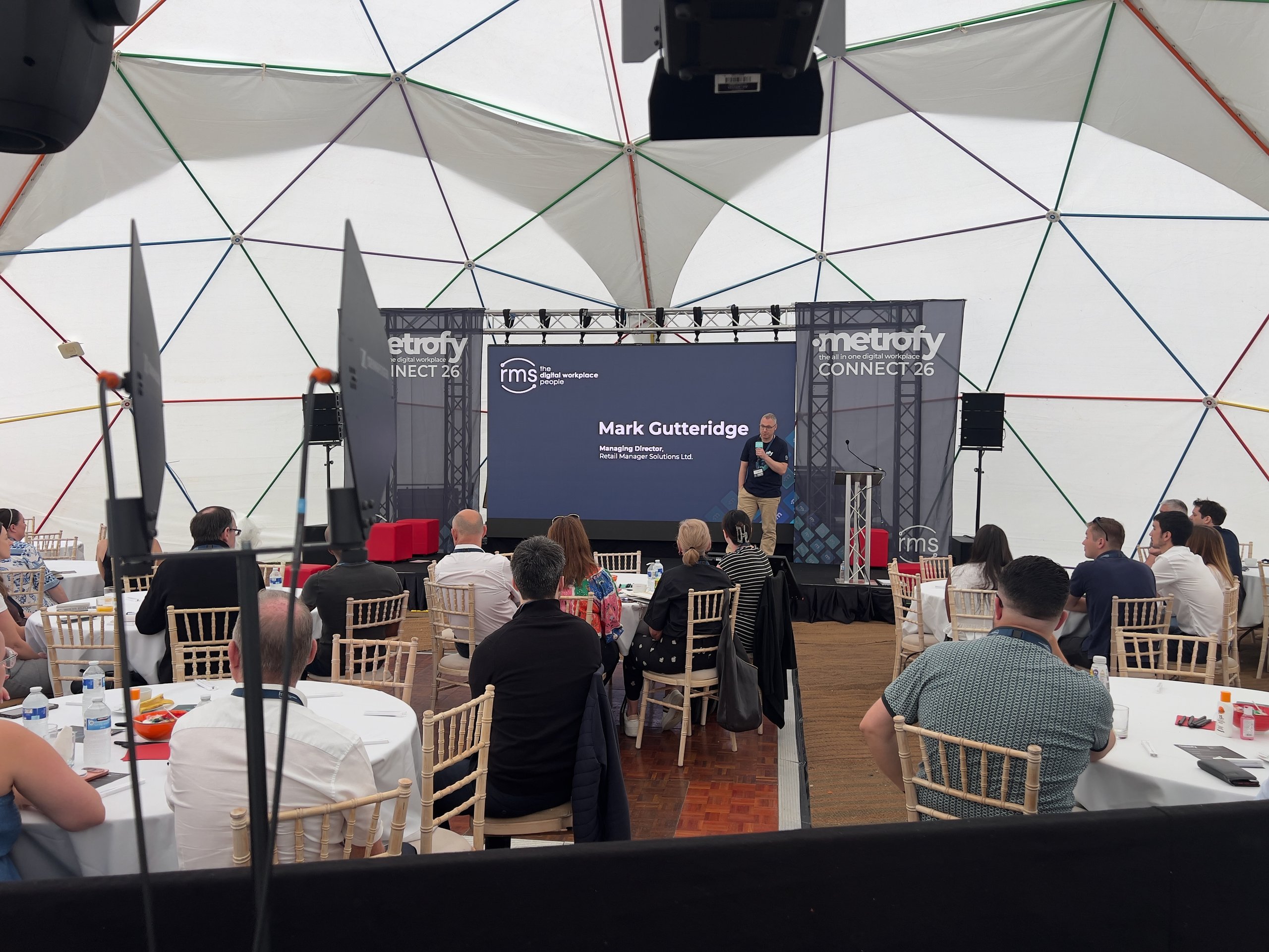 Speaker on stage at a corporate conference inside a dome, with attendees seated at round tables, professional AV setup visible.