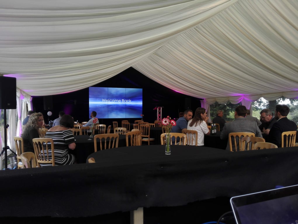 Guests seated at round tables in a draped marquee with a large video screen displaying Welcome Back, ready for a corporate event.