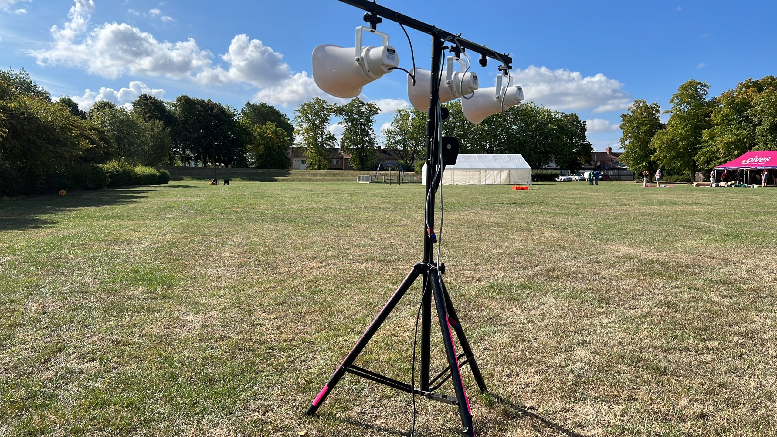 Outdoor event setup with stage lighting equipment in a grassy field, tents in the background, and people preparing for a gathering.