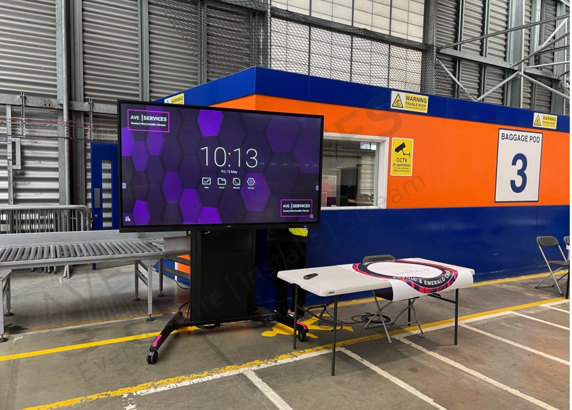 Large display screen setup in an industrial venue, near baggage area 3, with AV equipment and table for event production.