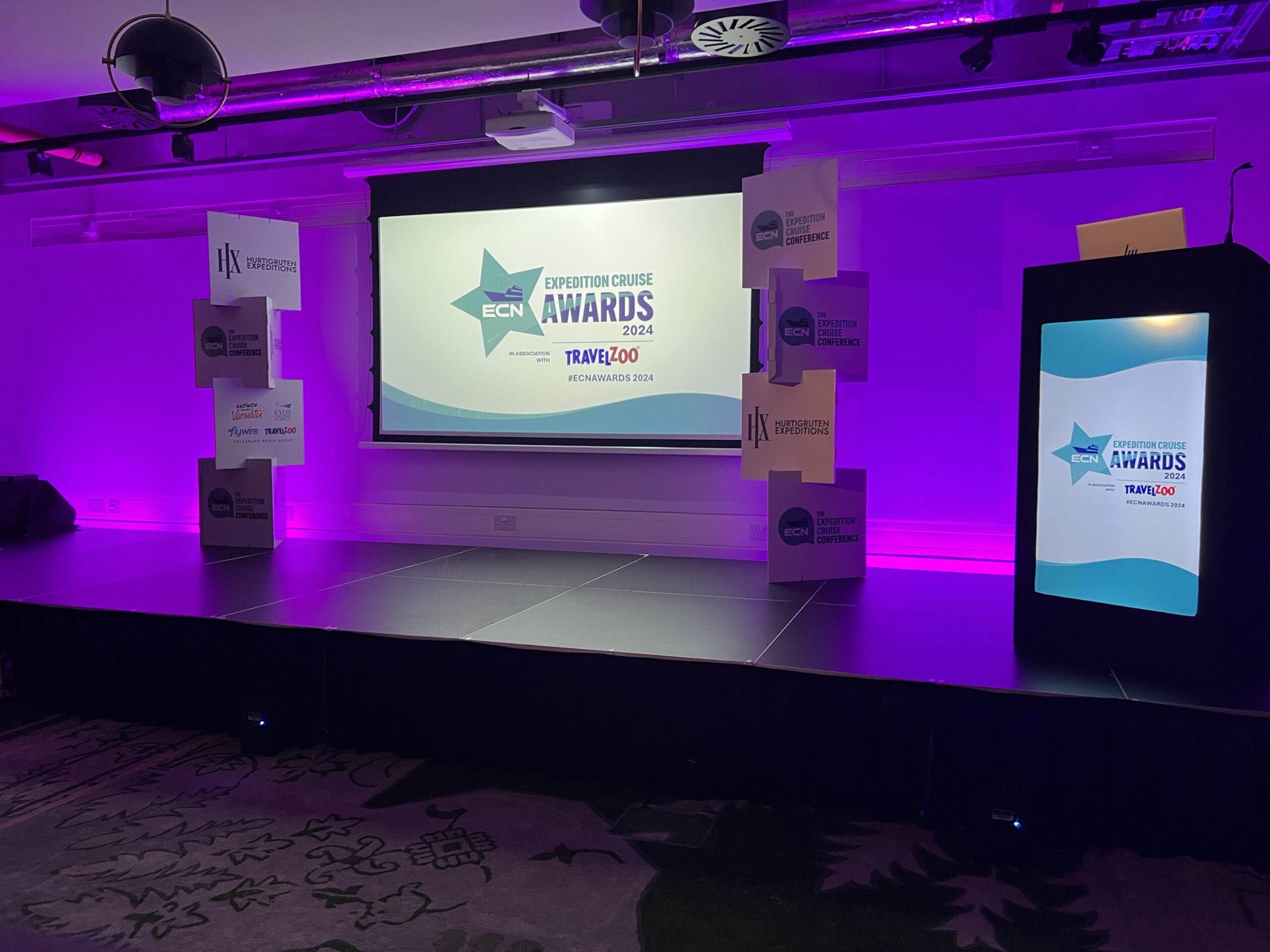 Stage setup for Expedition Cruise Awards 2024 with purple lighting, a large display screen, and podium in an event venue.