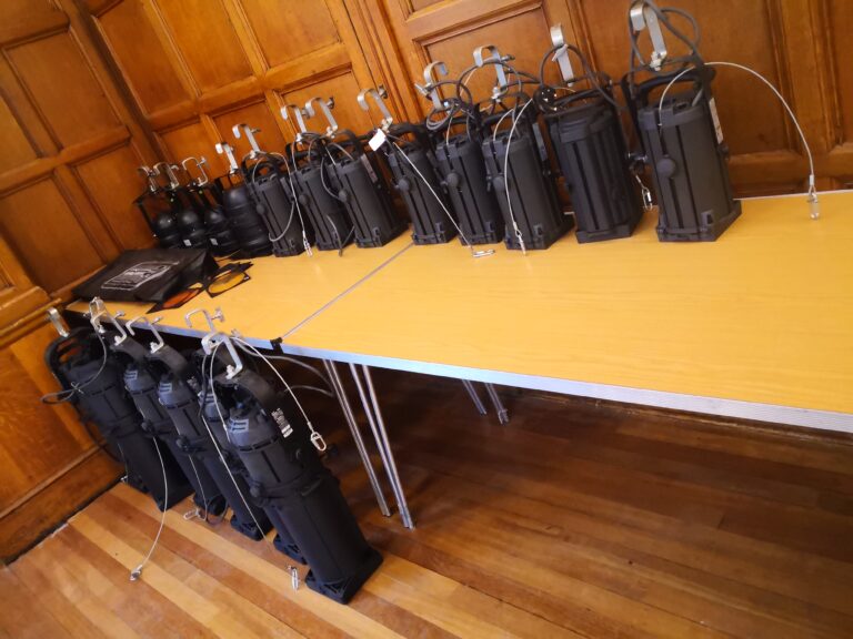 Stage lighting equipment organized on wooden tables, ready for event setup in a paneled venue room.