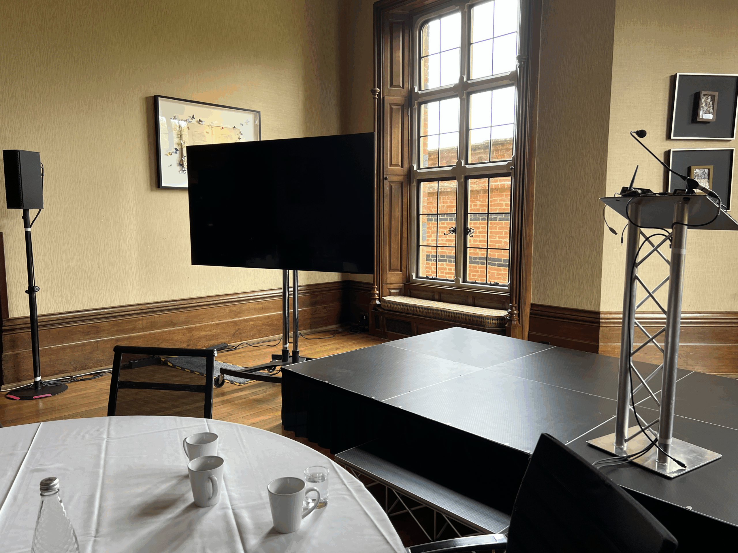 Staging setup in a conference room with a podium, large screen, and sound equipment, near a window.
