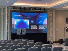 Conference room setup with a large LED screen displaying graphics, truss framework, seating for attendees, and AV equipment.