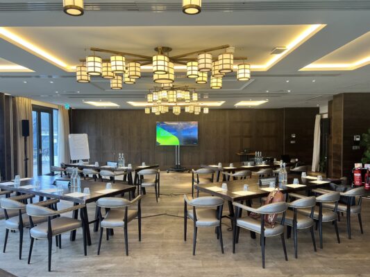 Conference room setup with staging, chairs, water bottles, flip chart, and large display screen for a corporate meeting.