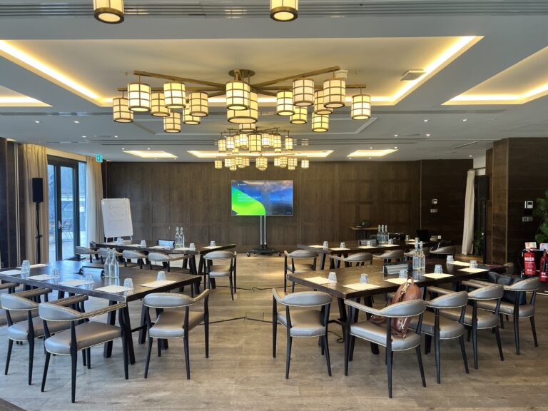 Conference room setup with staging, chairs, water bottles, flip chart, and large display screen for a corporate meeting.