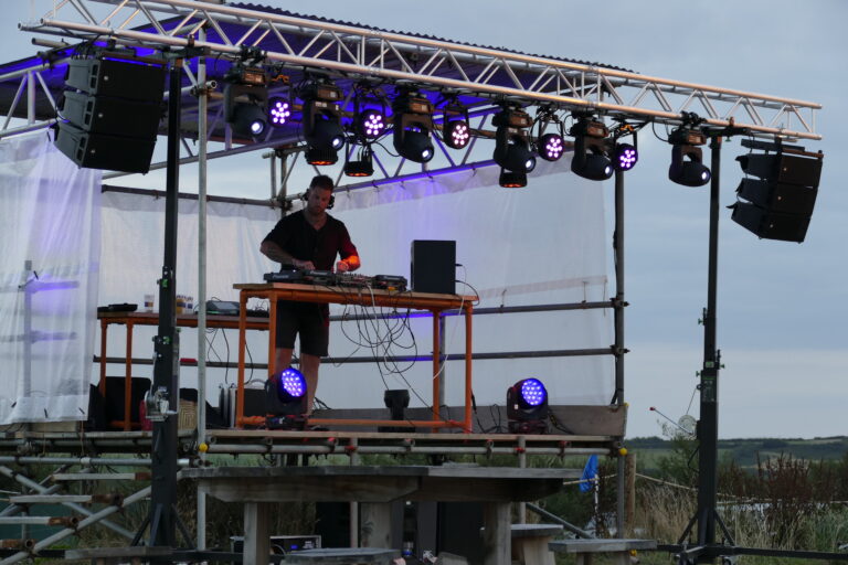 DJ performing on an outdoor stage with professional lighting and sound equipment, set against a scenic backdrop.