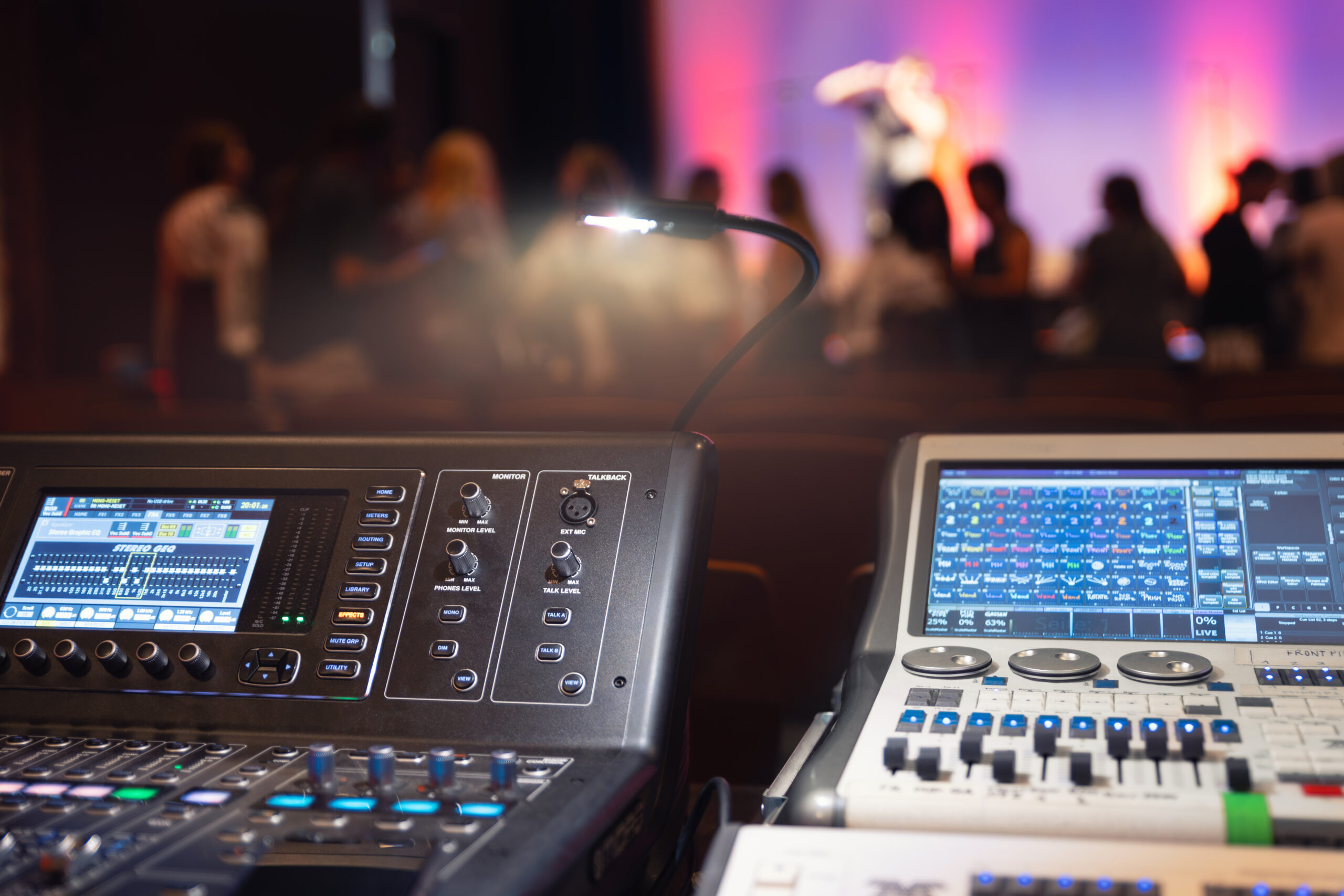 Sound and lighting control panel at a live event, with blurred audience and stage in the background.