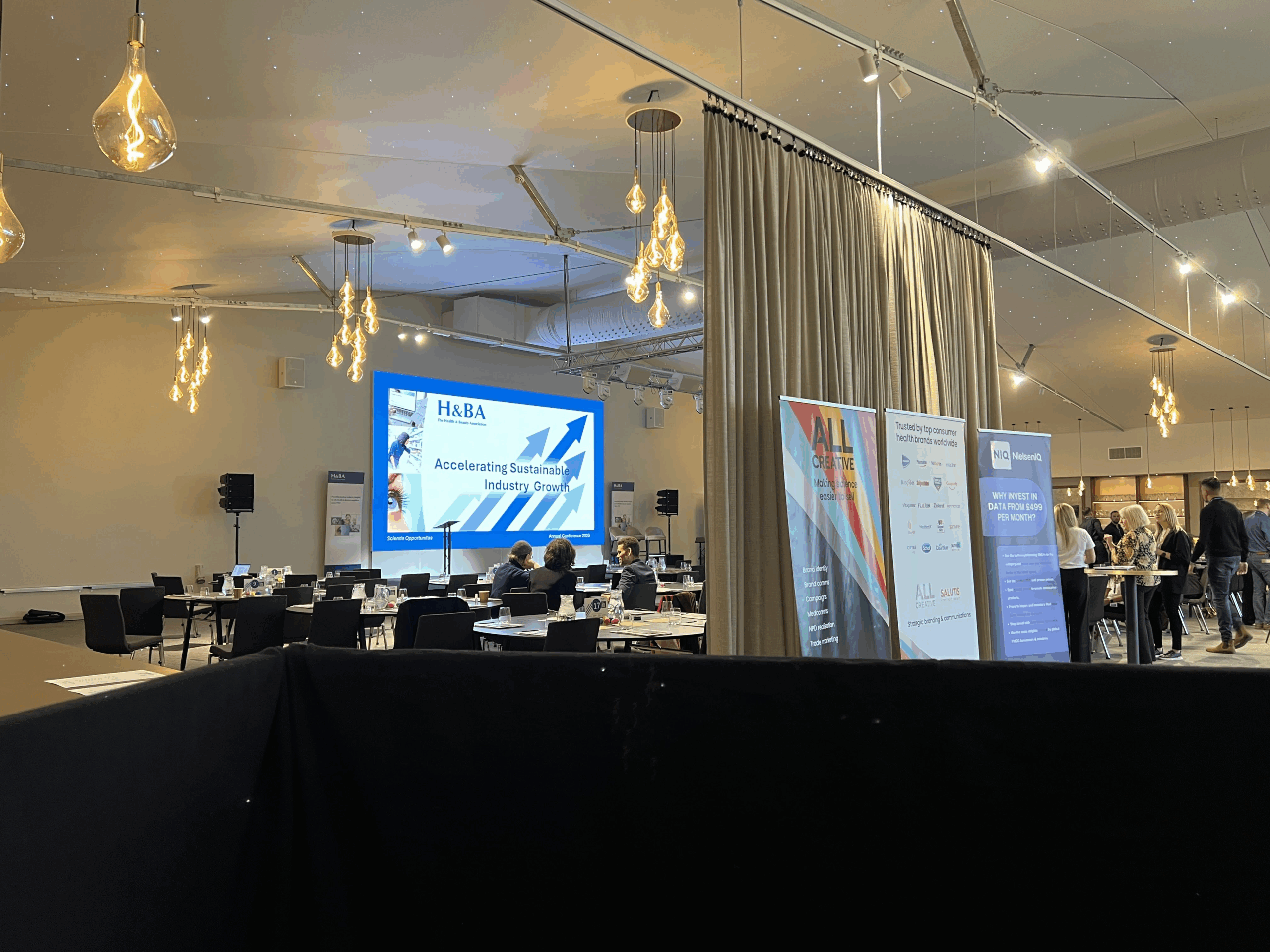 Corporate conference setup with staging, screen projection, and lighting. Attendees gather around tables with promotional banners visible.