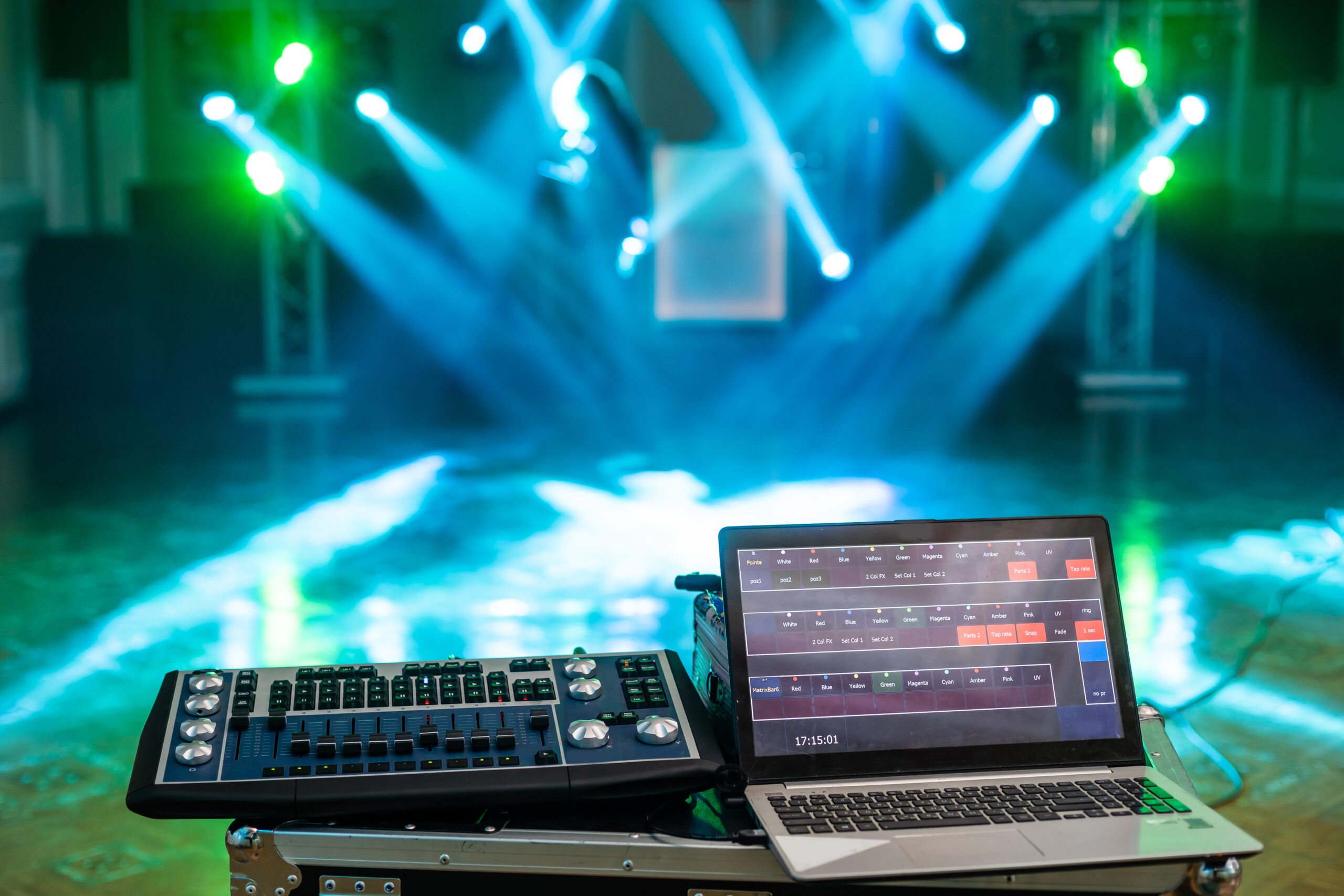 Lighting control console and laptop setup for event staging with vibrant blue and green stage lighting effects.