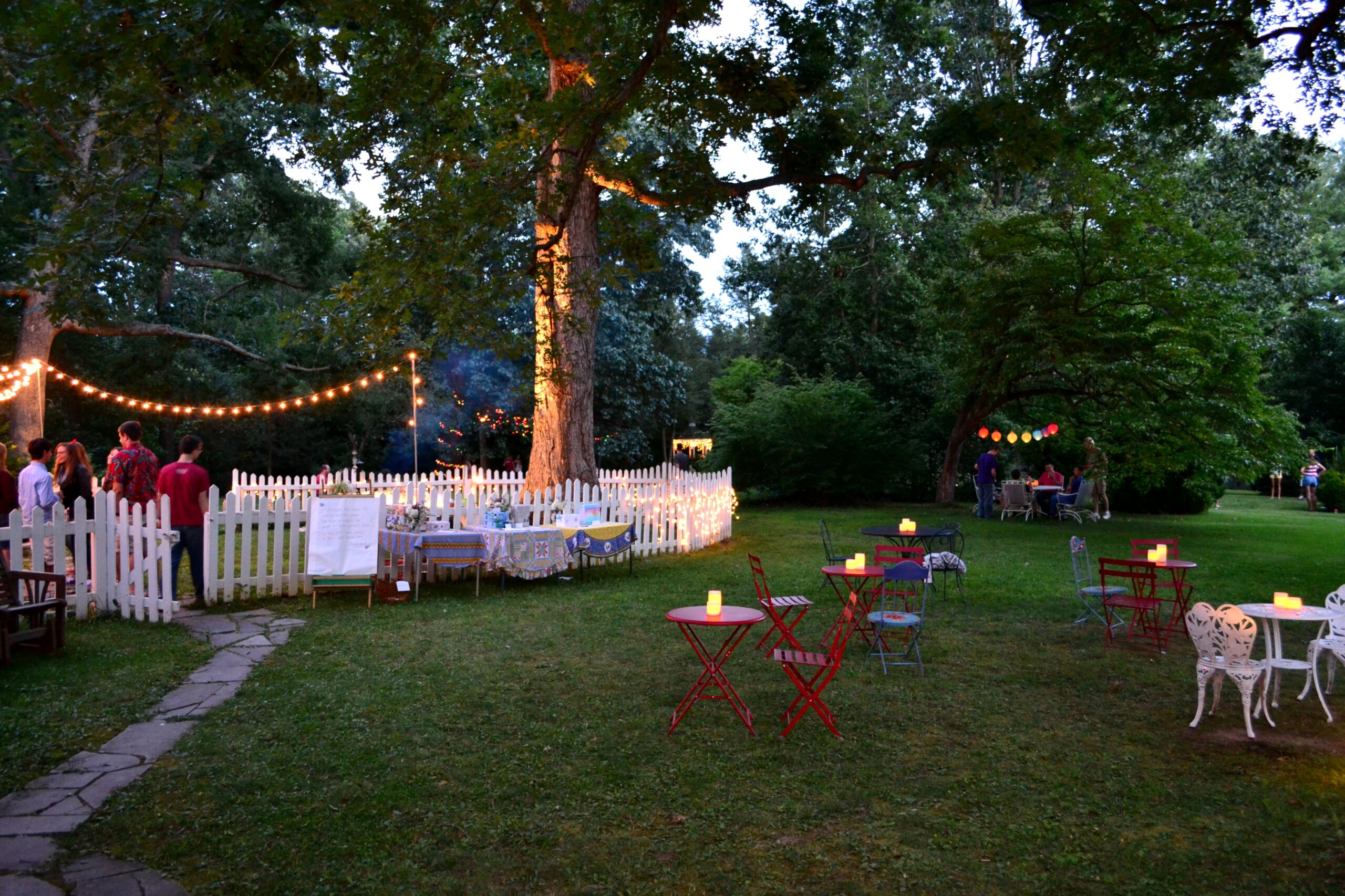 Outdoor garden party with string lighting, tables with candles, and guests gathering near a white picket fence.